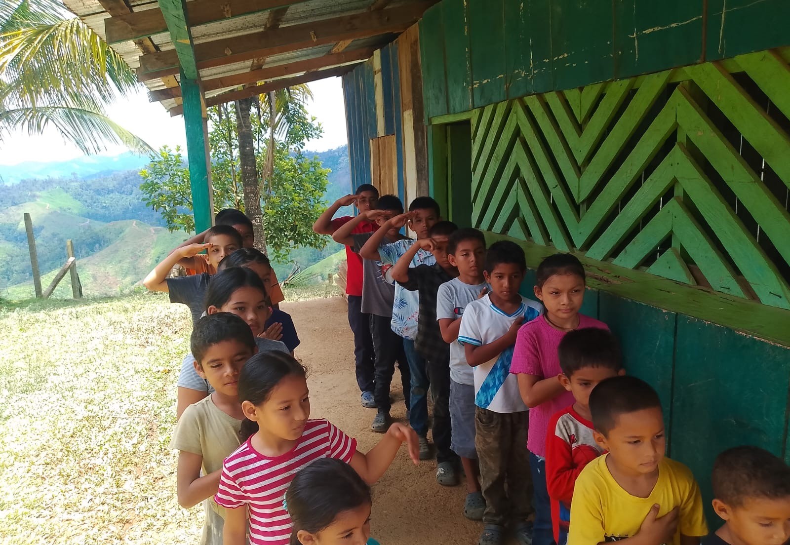 Children lined up on a path beside a green wooden school building, with green mountains in the distance